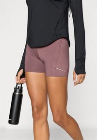 Shorts de sport courts en tissu mauve atténué, avec des coutures latérales et un détail de petits points bleu clair près de l'ourlet, associés à un haut noir.