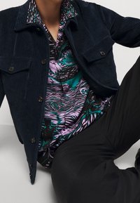 Veste en velours côtelé bleu marine avec boutons noirs et deux poches poitrine, portée sur une chemise à manches courtes colorée aux motifs abstraits violets et bleu sarcelle.