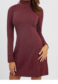 Femme portant une robe col roulé bordeaux à manches longues, côtelée et scintillante, posant avec une main sur la hanche et l'autre le long du corps.