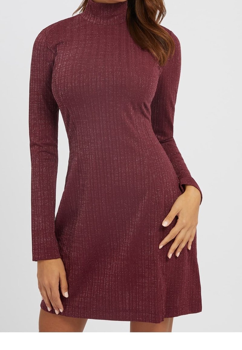 Femme portant une robe col roulé bordeaux à manches longues, côtelée et scintillante, posant avec une main sur la hanche et l'autre le long du corps.