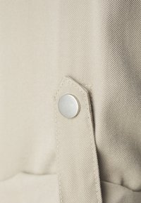 Beige fabric with a fine texture features a triangular flap and a round silver button. The detail showcases smooth stitching.