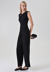 Combinaison noire avec un corsage structuré, un décolleté asymétrique et un pantalon à jambes larges. Présente un accent doré et est associée à des ballerines noires.