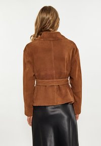 Giacca in suede marrone con vita regolabile, ampio colletto e design peplum. Texture liscia con dettagli di cucitura visibili.