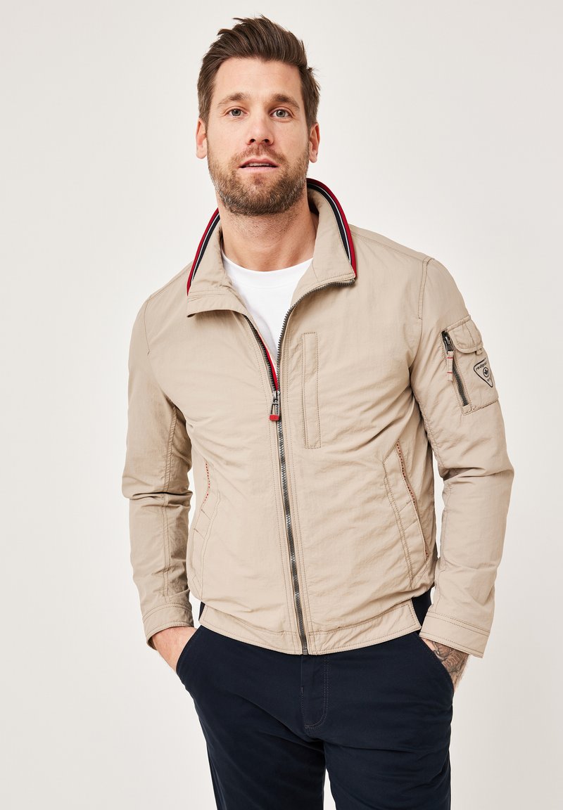 Beige jacket with a high collar, red and blue striped accents, front zipper, and side pockets. Worn over a white shirt, paired with navy pants.