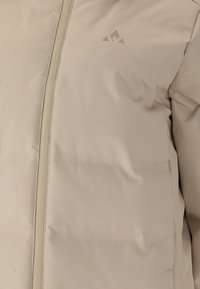 Beige kuplaruotsin takki vetoketjulla, jossa on sileä pinta ja minimalismi muotoilu. Pien logo sijaitsee vasemmassa yläkulmassa.