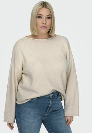 Crème gekleurde trui met een brede halslijn en lange mouwen, gecombineerd met blauwe denim jeans. De stof heeft een licht onregelmatig oppervlak.
