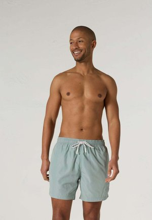 Hombre sonriente sin camiseta con pantalones cortos de baño azul claro con cordón blanco, de pie frente a un fondo liso y claro.