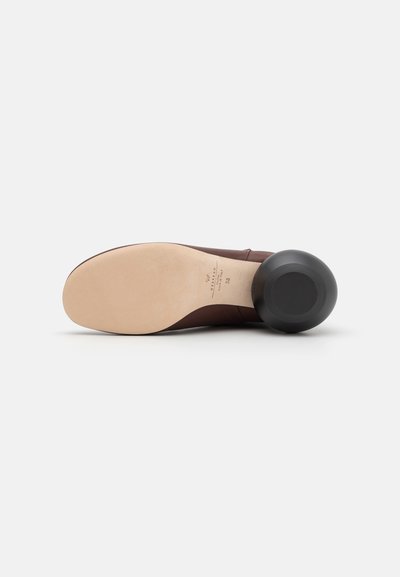 Chaussure marron avec un dessus en cuir lisse et un talon noir arrondi. La semelle est beige et plate, avec des détails de marque discrets.