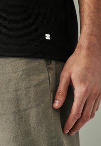 Maglietta nera a costine con un piccolo logo argento all'orlo, abbinata a pantaloni leggeri color khaki. Focus sulla texture del tessuto e sui dettagli del design.