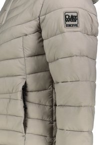 Beige Steppjacke mit strukturiertem Design, ausgestattet mit einer seitlichen Tasche mit Reißverschluss und einem auffälligen Markenpatch auf der Brust. Glattes, glänzendes Material.