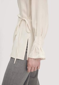 Blusa beige chiaro realizzata in tessuto morbido, con vita legata, maniche svasate e spacchi laterali, abbinata a pantaloni grigi con texture.