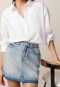 Chemise blanche à boutons avec manches retroussées, associée à une jupe en denim bleu clair. La jupe présente un ourlet effiloché et cinq poches.