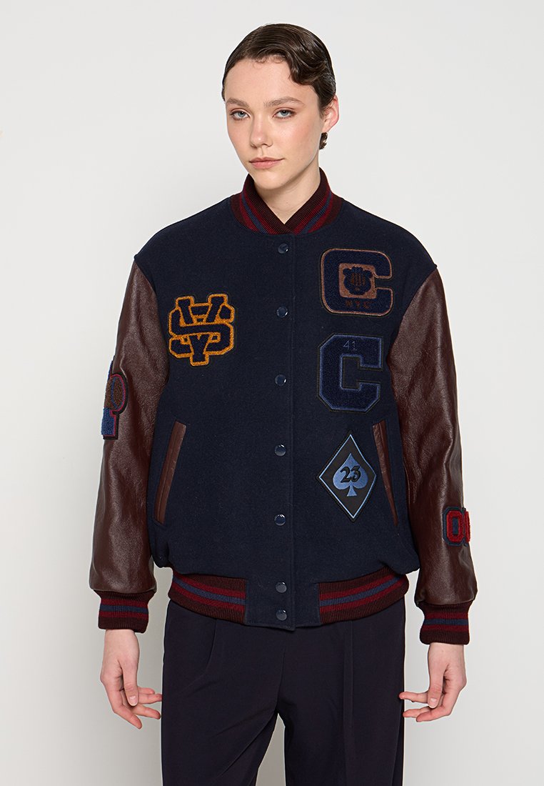 Giacca varsity in lana navy con maniche in pelle marrone, caratterizzata da toppe multicolori, colletto a coste e strisce colorate sui polsini e sull'orlo.