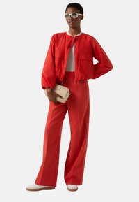 Rød bomberjakke med elastiske manchetter, matchende vidde bukser og hvide sko. Modellen holder en cremefarvet clutch. Runde solbriller fuldender looket.