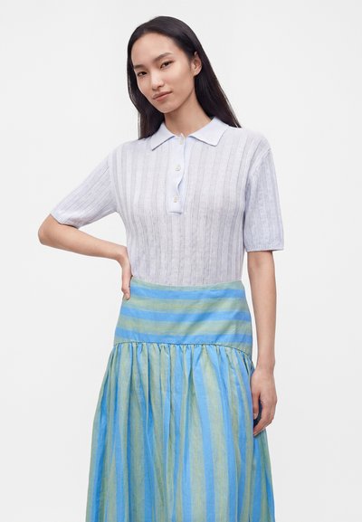 Jeune femme aux longs cheveux noirs porte un polo en maille légère à manches courtes et une jupe rayée bleue et verte, debout avec la main sur la hanche.