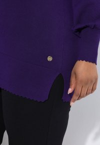 Maglione viola con orlo scallop e polsini a costine, con un piccolo tag logo dorato. Abbinato a leggings neri. Texture di maglia liscia.