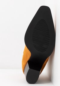 Botte pointue en daim marron avec semelle en caoutchouc noire et talon bloc, inclinée contre un fond d'angle blanc.