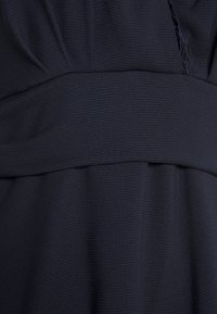 Abito navy con tessuto a trama, caratterizzato da un vita aderente e un design drappeggiato. Dettaglio in pizzo sottile lungo il decollété.