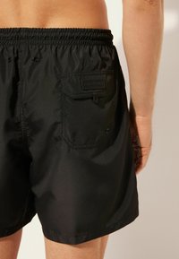 Bañador negro hecho de tela ligera, con cinturilla elástica, bolsillo trasero y pequeño detalle de logo. Textura suave.