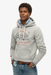 Lysegrå hættetrøje med en frontlomme, snoretræk og trykt logo. Bæret over en denimskjorte og kombineret med blå jeans og et brunt bælte.
