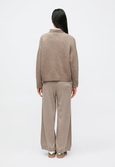 Pikkade tume juustega naine seisab seljaga vaataja poole, kandes lahtist taupe tooni kampsunit, laiade jalgadega kootud pükse ja kahetoonilisi tossusid valgel taustal.