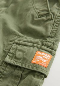 Maslinasta tkanina s teksturiranim završetkom, koja sadrži istaknutu narančastu oznaku s natpisom "SUPER DRY" i detaljima šivanja na džepu.