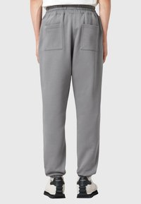 Vedere din spate a unei persoane care poartă pantaloni de trening gri cu talie elastică și două buzunare la spate, asortați cu adidași albaștri și negri.