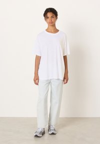 T-shirt bianca a maniche corte con vestibilità rilassata, abbinata a jeans a gamba larga azzurri chiari. Il modello indossa sneakers grigie, in piedi su un pavimento neutro.