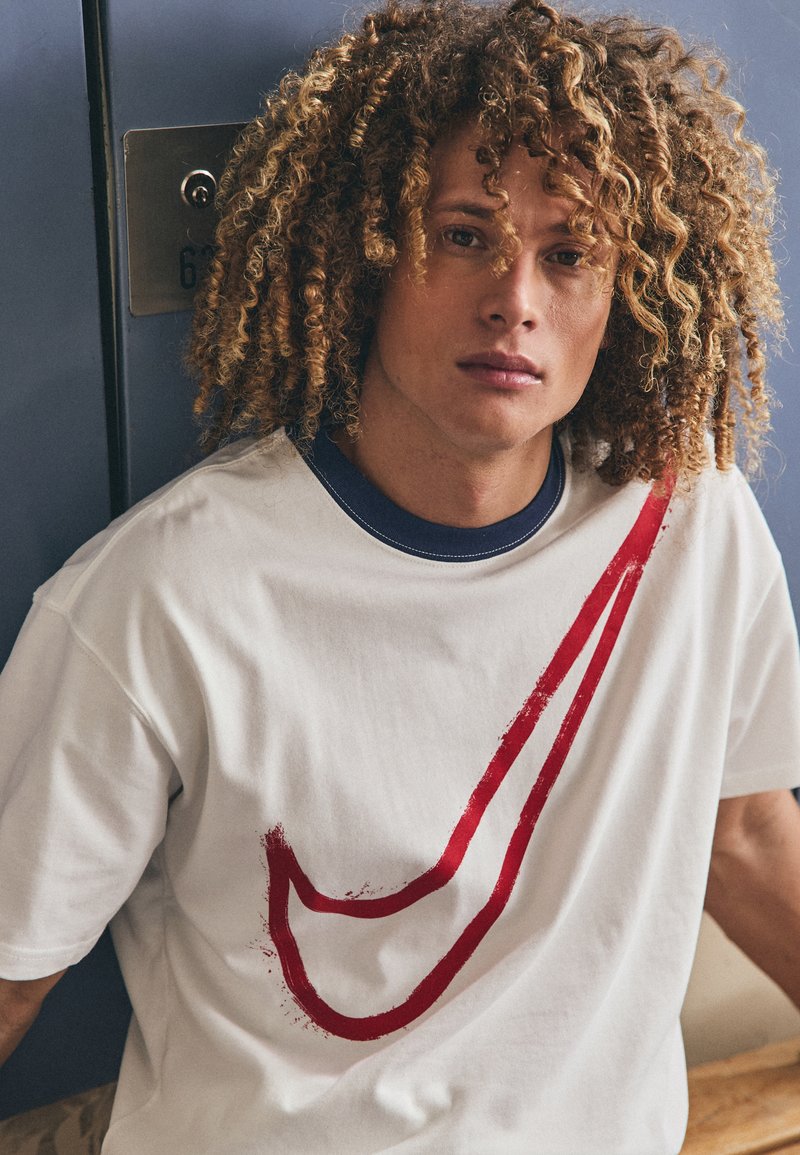 Camiseta de algodón blanca con un gran diseño de swoosh rojo, cuello redondo en color marino y mangas cortas. La textura parece suave y ligera.