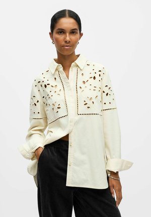 Cremfarbene Bluse mit ausgeschnittenen Blumen- und geometrischen Mustern. Mit Kragen, langen Ärmeln und lässiger Passform. Kombiniert mit dunkelbraunen Cordhosen.