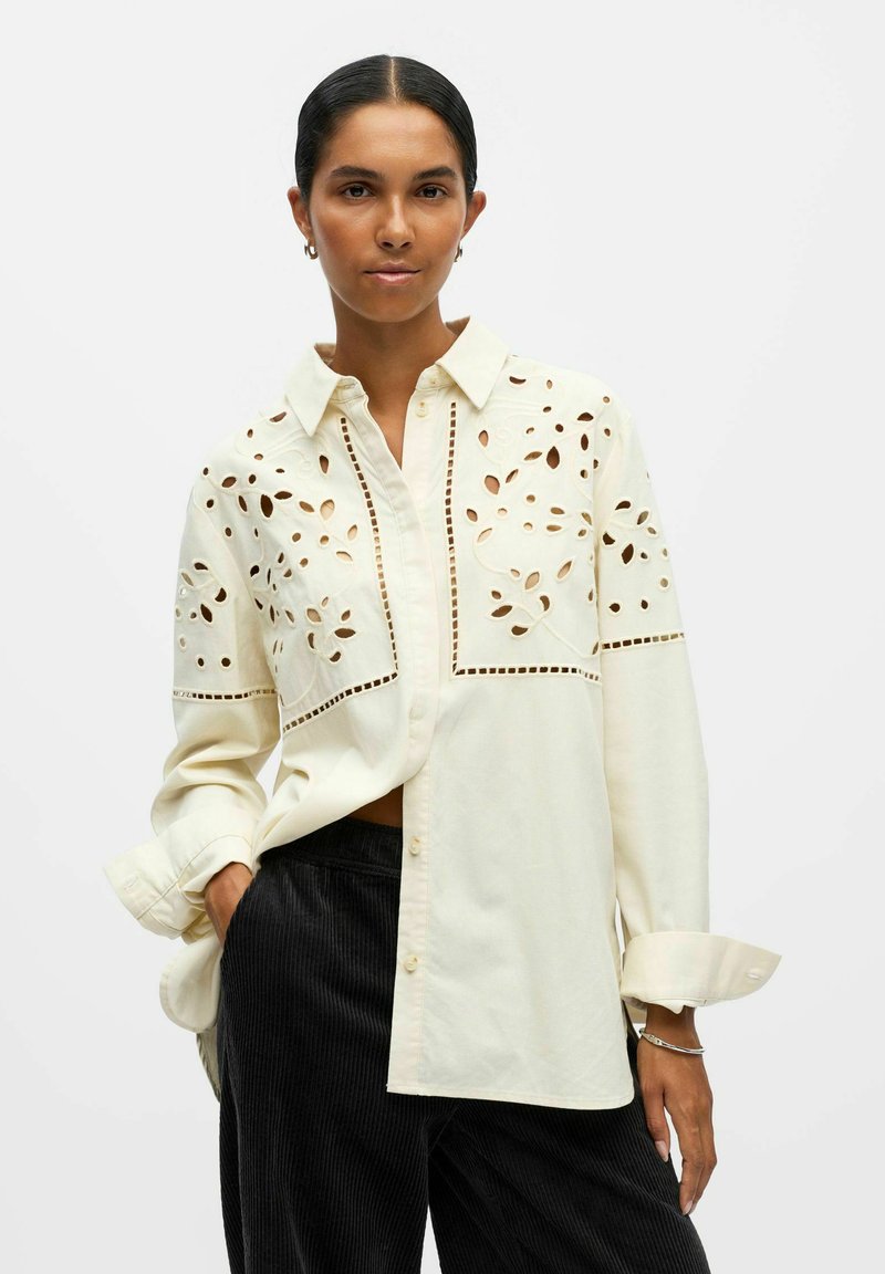 Cremfarbene Bluse mit ausgeschnittenen Blumen- und geometrischen Mustern. Mit Kragen, langen Ärmeln und lässiger Passform. Kombiniert mit dunkelbraunen Cordhosen.