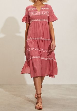 Roze jurk tot de knie met een v-hals, korte uitlopende mouwen en bloemenbroderie langs horizontale banden. Lichtgewicht stof met een vloeiende silhouet.