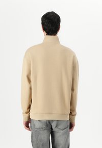 Sweatshirt beige con alto colletto, vestibilità rilassata, polsini e orlo a coste, realizzato in tessuto morbido. Indossato con jeans grigi, visto da dietro.