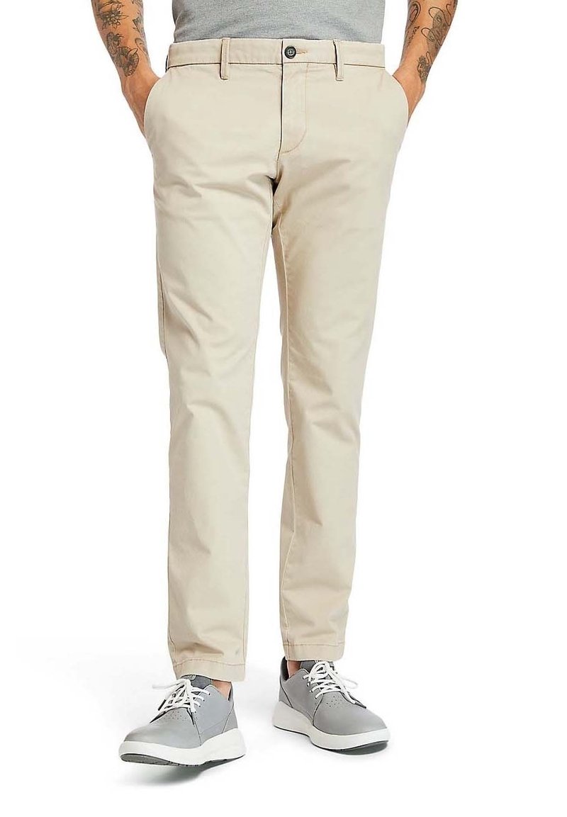 Pantalon chino en sergé beige à coupe slim, avec fermeture à boutons sur le devant, poches latérales et texture lisse, associé à des baskets grises.