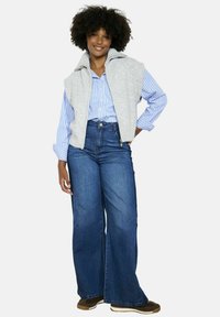 Pelēks bezpieks ar augstu apkakli virs zila un balta svītrainā krekla ar pogām, savienots ar augsto vidukli un plašiem ziliem džinsiem.