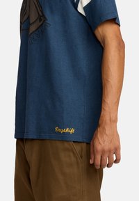 T-shirt bleu marine à manches courtes avec un design graphique, arborant un "Dayshift" brodé en doré près de l'ourlet. Matière en coton, coupe décontractée.