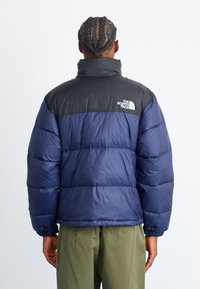 Marineblå og sort puffjakke med høj krave, der har et logo på øverste ryg og ribbede manchetter, lavet af skinnende, isoleret materiale.