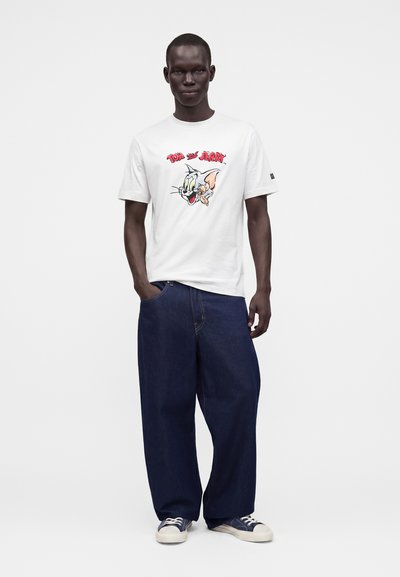 Homme portant un t-shirt blanc avec un motif Tom et Jerry, un jean bleu foncé ample et des baskets bleues, debout avec une main dans la poche.