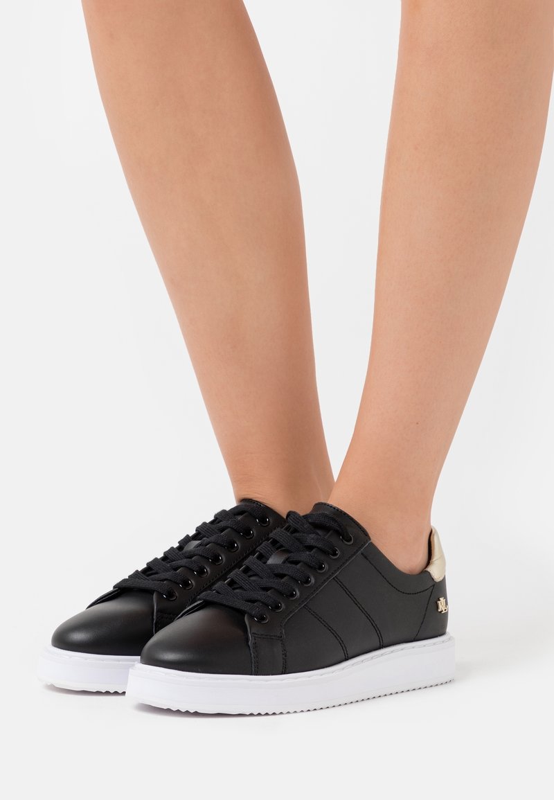 Baskets basses noires avec semelles blanches portées pieds nus, montrant des vues de côté et de face sur un fond clair uni.