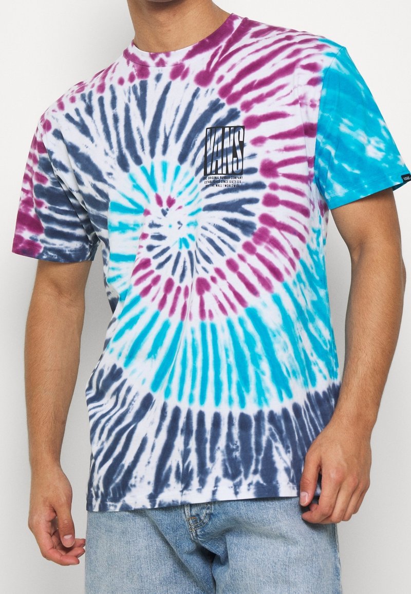 Homme portant un t-shirt Vans tie-dye multicolore avec un jean bleu, main reposant près de la poche, sur un fond uni.