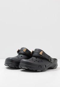 Paire de chaussures noires style sabot à enfiler, avec des trous de ventilation et des brides ajustables, posées sur une surface blanche contre un fond uni.