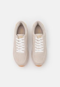 Beżowe zamszowe sneakersy z białymi sznurówkami, zaokrąglonym noskiem i jasnobrązową gumową podeszwą. Charakteryzują się minimalistycznym designem i wytłoczonym logo na języku.