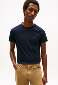 Marineblå t-shirt med korte ærmer og rund halsudskæring, med en lille logoaccent; sat sammen med lyse beige bukser.