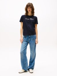 T-shirt en coton bleu marine avec "Tommy Hilfiger" en écriture blanche, associé à un jean large bleu clair et des chaussures blanches. Tenue simple et décontractée.
