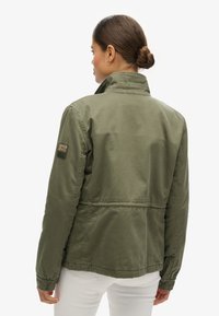 Veste en olive en coton, avec un col montant, des poignets élastiques et un patch logo sur la manche gauche. Coupe ajustée.