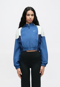 Veste Nike bleue recadrée avec des accents blancs et bleu marine, fermeture éclair à l'avant, col haut et poignets élastiques. Tissu lisse avec une coupe ajustée.