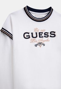 Witte biologische katoenen t-shirt met een marineblauwe en beige ribgebreide kraag. Voorzien van geborduurd "GUESS Los Angeles" logo en retro graphics op de voorkant.