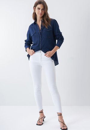 Chemise en denim bleu marine avec deux poches poitrine et fermeture à boutons, associée à un pantalon skinny blanc et des escarpins noirs à lanières.
