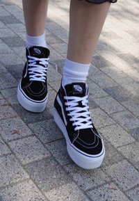 Zwarte hoge sneakers met witte veters en buitenzool. Suède bovenwerk met contrasterende stiksels en logo-patch op de tong. Draag met witte sokken.