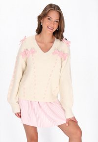 Cremfarbener Strickpullover mit V-Ausschnitt, versehen mit rosa Schleifen und strukturierten Maschendetails. Kombiniert mit einem hellrosa, gefalteten Rock.